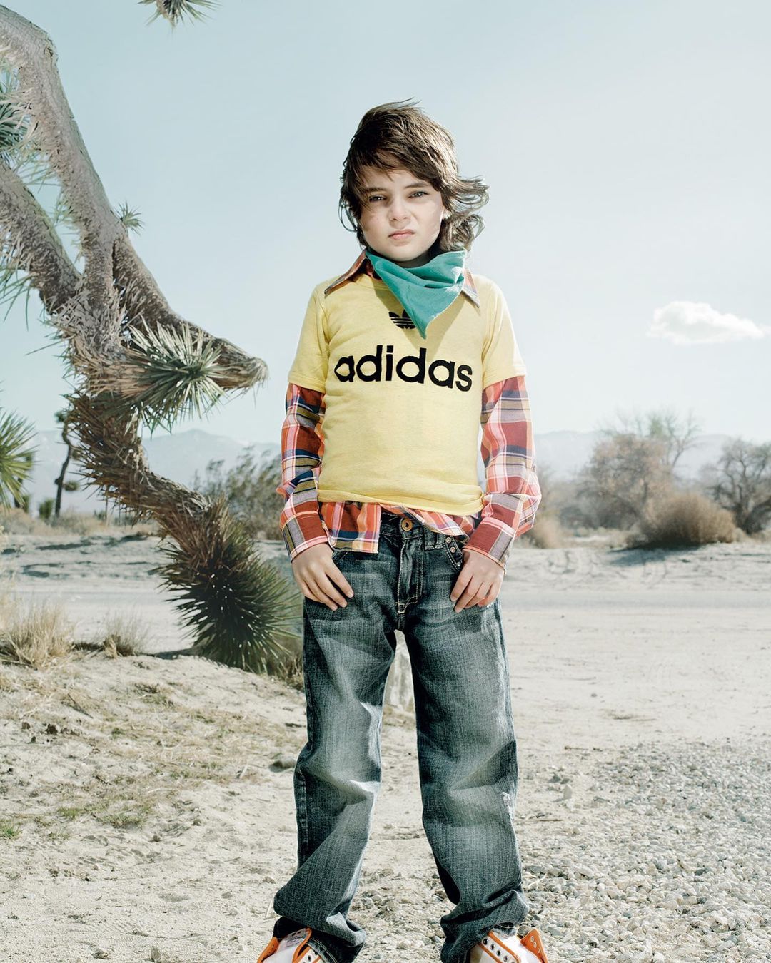 A boy in a yellow Adidas shirt, jeans, and a turquoise scarf poses in a desert landscape with arid plants under a clear sky.