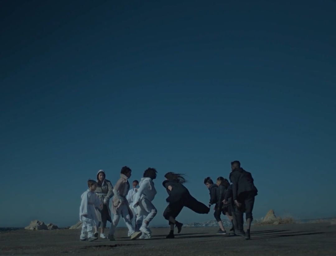 A group of people stand on a tan surface with an open blue sky in a video or photograph; some wear light clothes and some wear dark clothes.