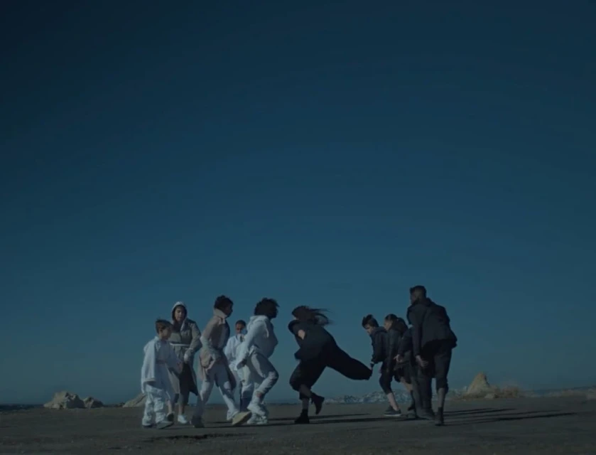 A group of people stand on a tan surface with an open blue sky in a video or photograph; some wear light clothes and some wear dark clothes.