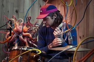 Musician with octopus art and colorful cables in a rustic music studio, wearing a cap, playing percussion instrument with drum kit.