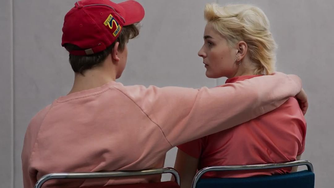 Back view of a couple sitting on chairs, embracing. The woman with blonde hair looks at the man in the red cap.
