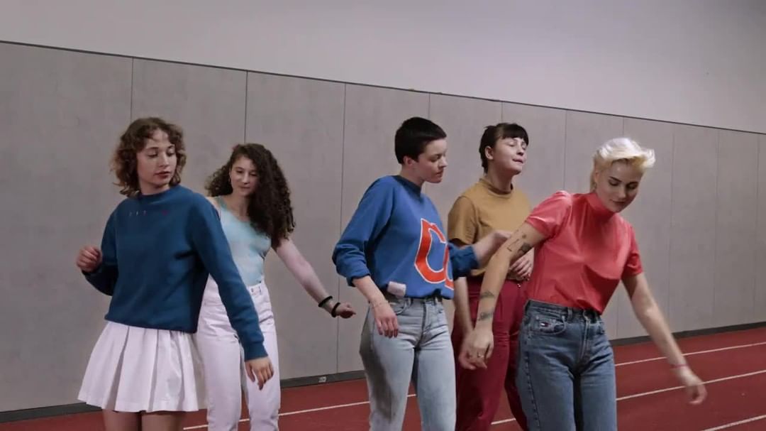 Five young adult women stand close to each other in an indoor track. They wear casual clothing, and there is natural lighting.