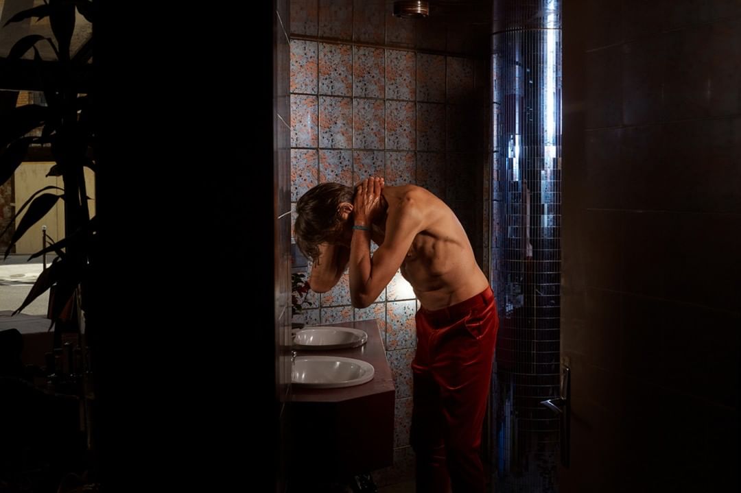 Man with bare torso washing his face in a bathroom with tiled walls, illuminated by an adjacent bright column of light.