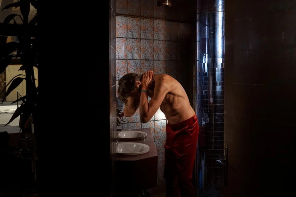 Man with bare torso washing his face in a bathroom with tiled walls, illuminated by an adjacent bright column of light.