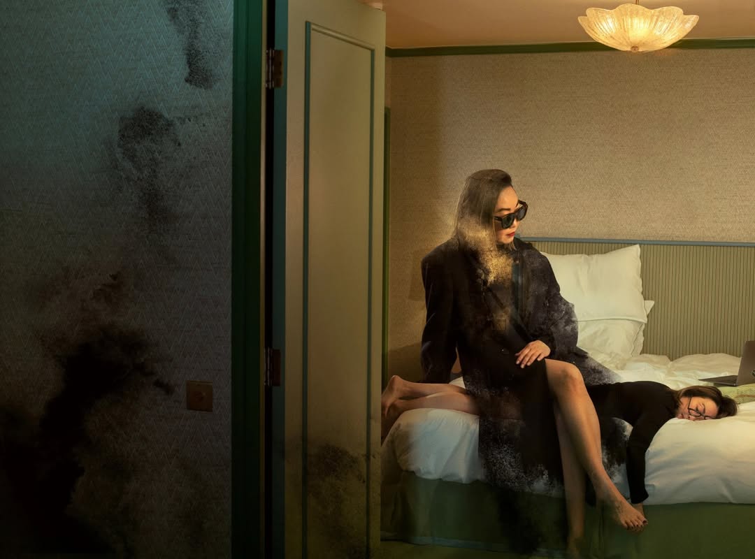 Two women in a hotel room, one sitting on the bed and the other lying across the bed with a laptop, in soft, diffused lighting.