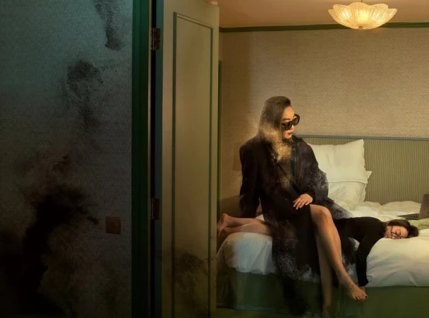 Two women in a hotel room, one sitting on the bed and the other lying across the bed with a laptop, in soft, diffused lighting.