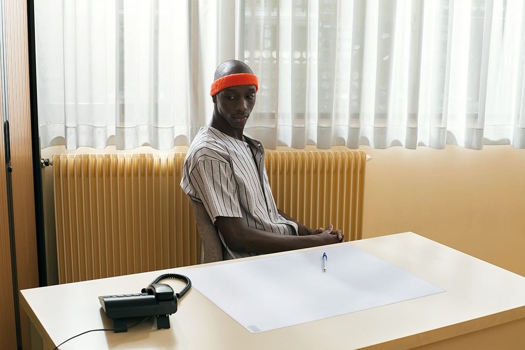 Portrait of a young, bald, Black man wearing a baseball shirt and orange headband. He sits at a desk in front of a window.