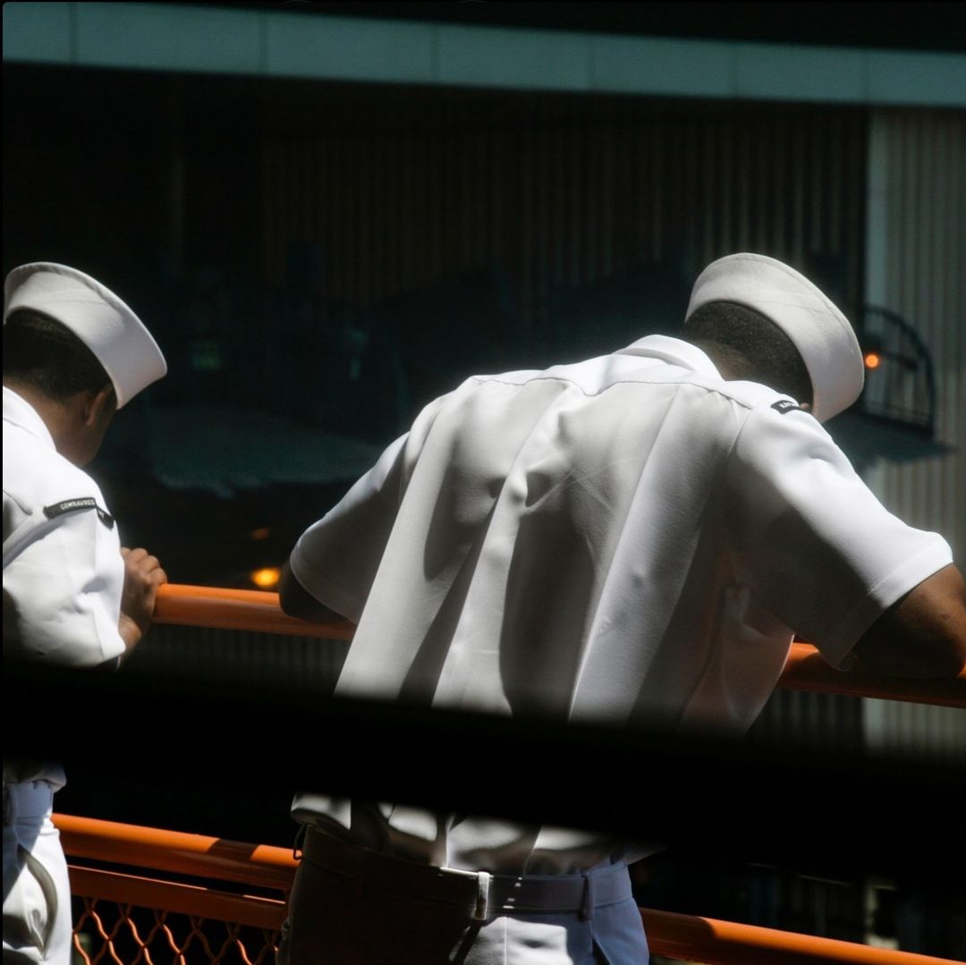 Two men in white uniforms stand on a deck or balcony with an orange railing, looking out at an obscured background. The scene is brightly lit.