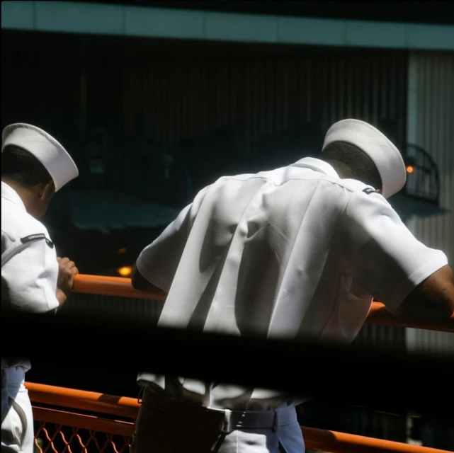 Two men in white uniforms stand on a deck or balcony with an orange railing, looking out at an obscured background. The scene is brightly lit.