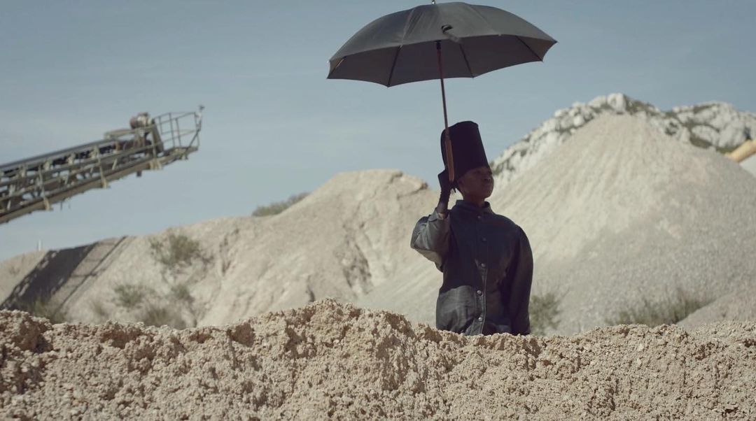 Person in formal attire and top hat holds a black umbrella against a backdrop of sandy dunes and quarry equipment under a blue sky.