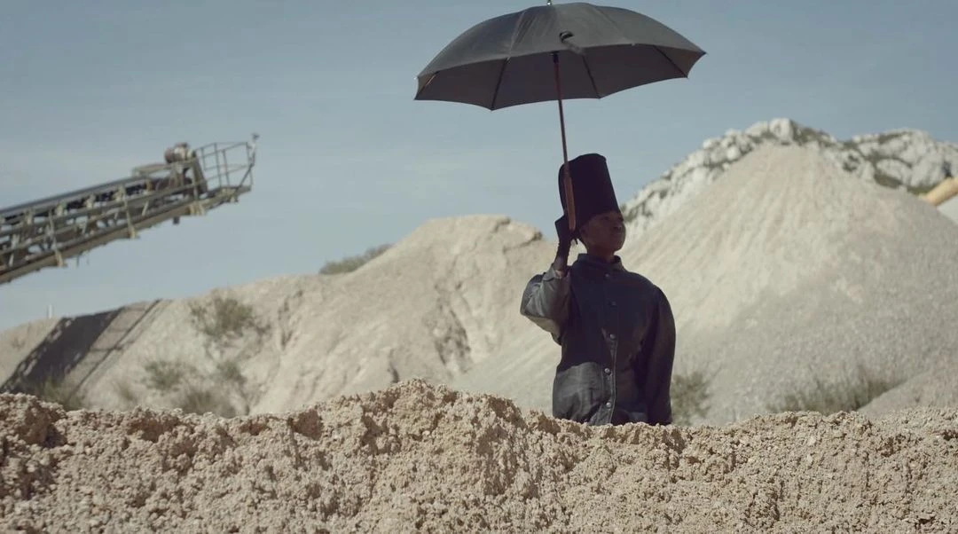 Person in formal attire and top hat holds a black umbrella against a backdrop of sandy dunes and quarry equipment under a blue sky.