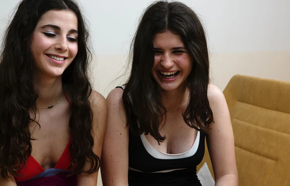 Two young women with long brown hair are closely seated indoors, one smiling slightly, the other laughing joyfully with open mouth.