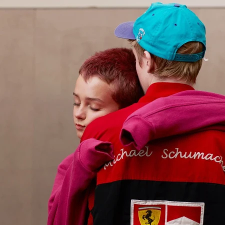 Two young people embrace indoors; one wears a red, black, and white racing jacket with logos, the other a pink hoodie and blue cap.