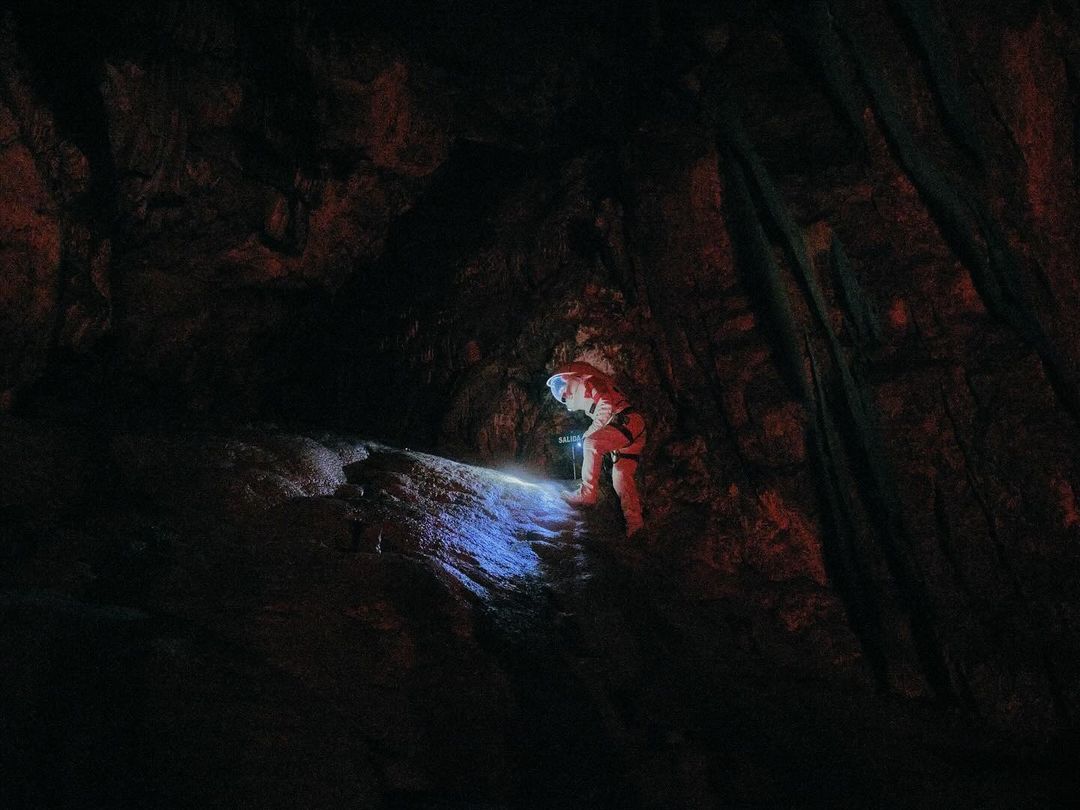 A person in a white jumpsuit and helmet explores a dark cave with a flashlight, illuminating the rugged rock formations inside.
