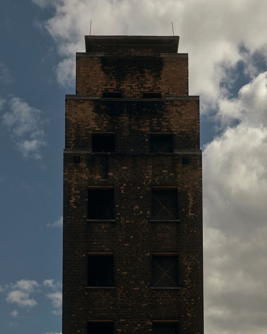 Exterior shot of a weathered brick building with visible water damage and boarded windows against a cloudy sky. Architecture photography.