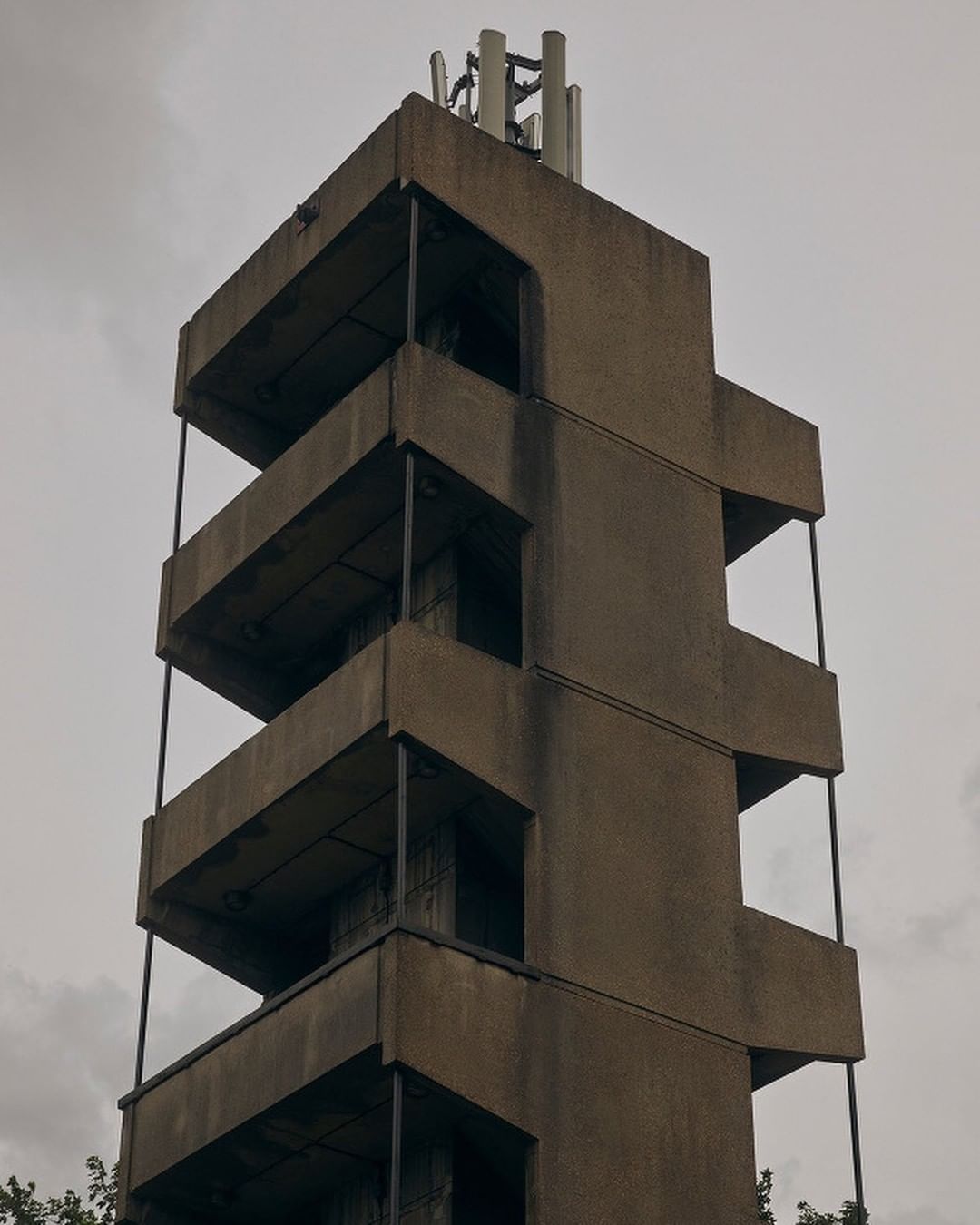 An exterior daytime shot features a tall, brutalist architectural structure with a symmetrical form and protruding balconies against a cloudy sky.