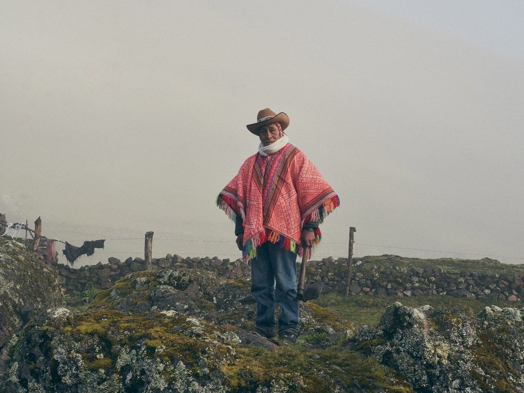 Adult male stands on a rocky hilltop wearing a red poncho and hat, holding an axe in a rural setting with a stone fence.