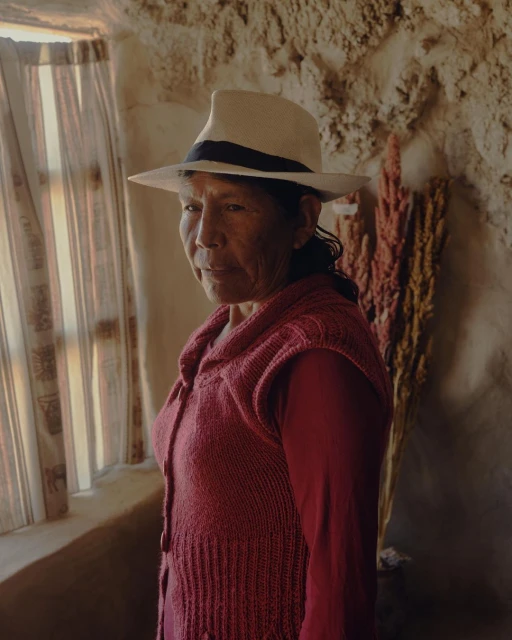An elderly woman with dark hair and a fair hat poses in a red sweater, inside a rustic room with a window and natural light.