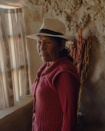 An elderly woman with dark hair and a fair hat poses in a red sweater, inside a rustic room with a window and natural light.