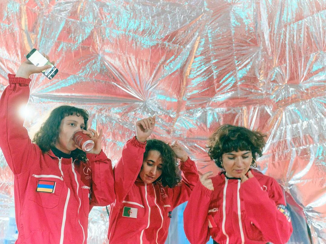 Three people in red jumpsuits pose against a silver background, one holding a phone, another drinking, creating a fun, dynamic scene.