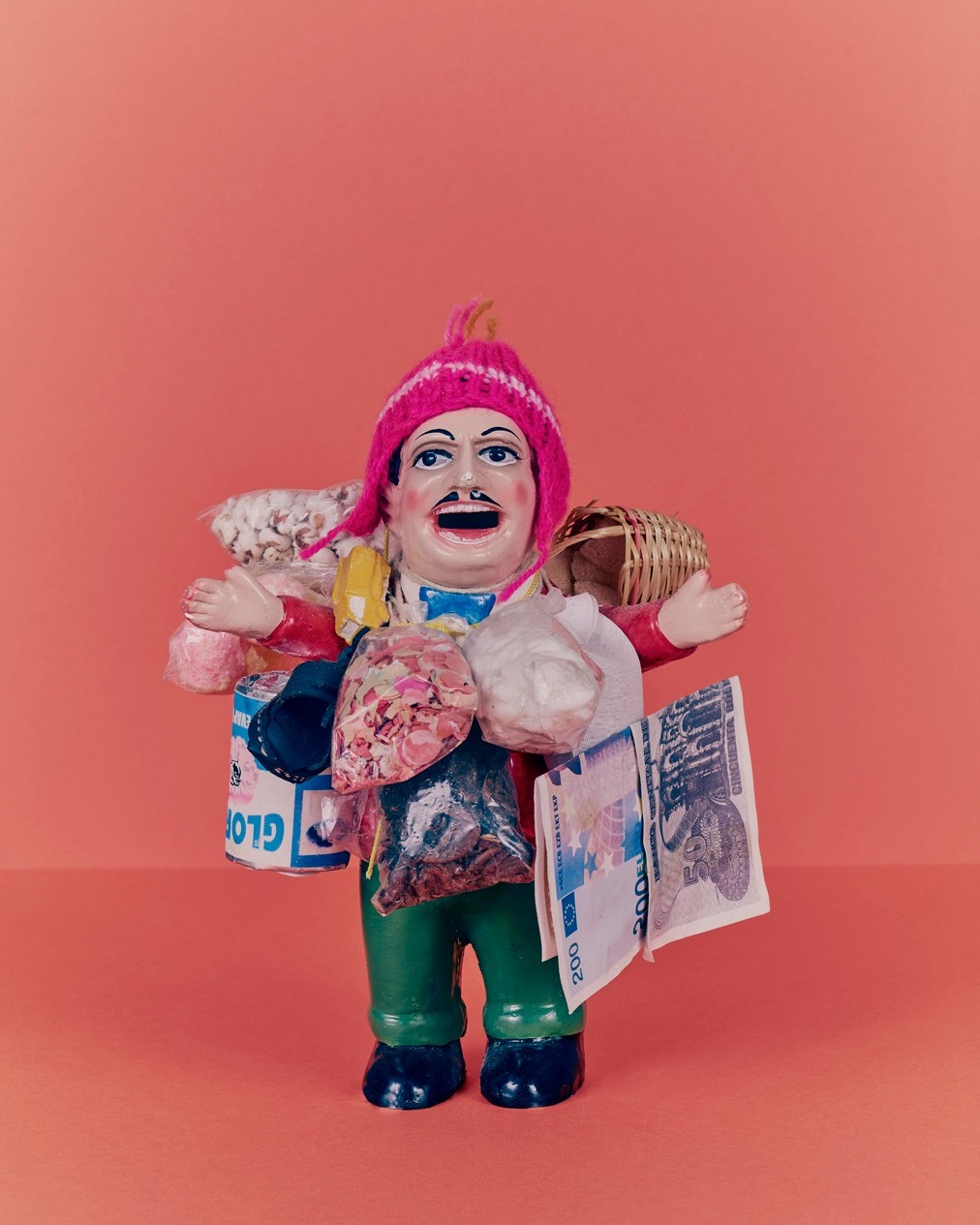 Figurine of a man wearing a pink hat carrying goods, including a basket, bags of food, and currency, against a salmon background.