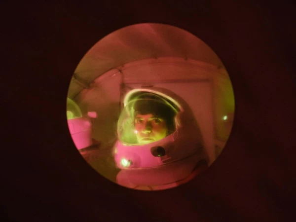 A person in an astronaut suit and helmet peers out through a round window in a dimly lit space station-like interior, illuminated in red and green.