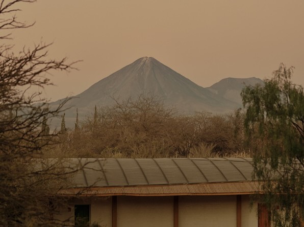 Landscape view of a large grey mountain peak framed by arid trees, brush, and the corner of a building with a solar panel roof.
