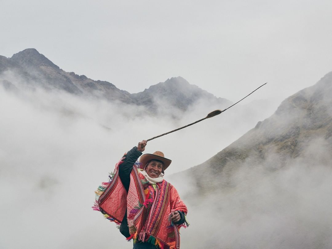 Adult male in traditional Andean clothing standing in a mountain landscape with clouds, wearing a hat, throwing a boleadoras, and smiling.