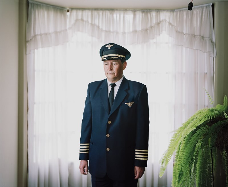 A uniformed senior man stands indoors near a window with white curtains and a potted plant. The subject is centered in the frame.