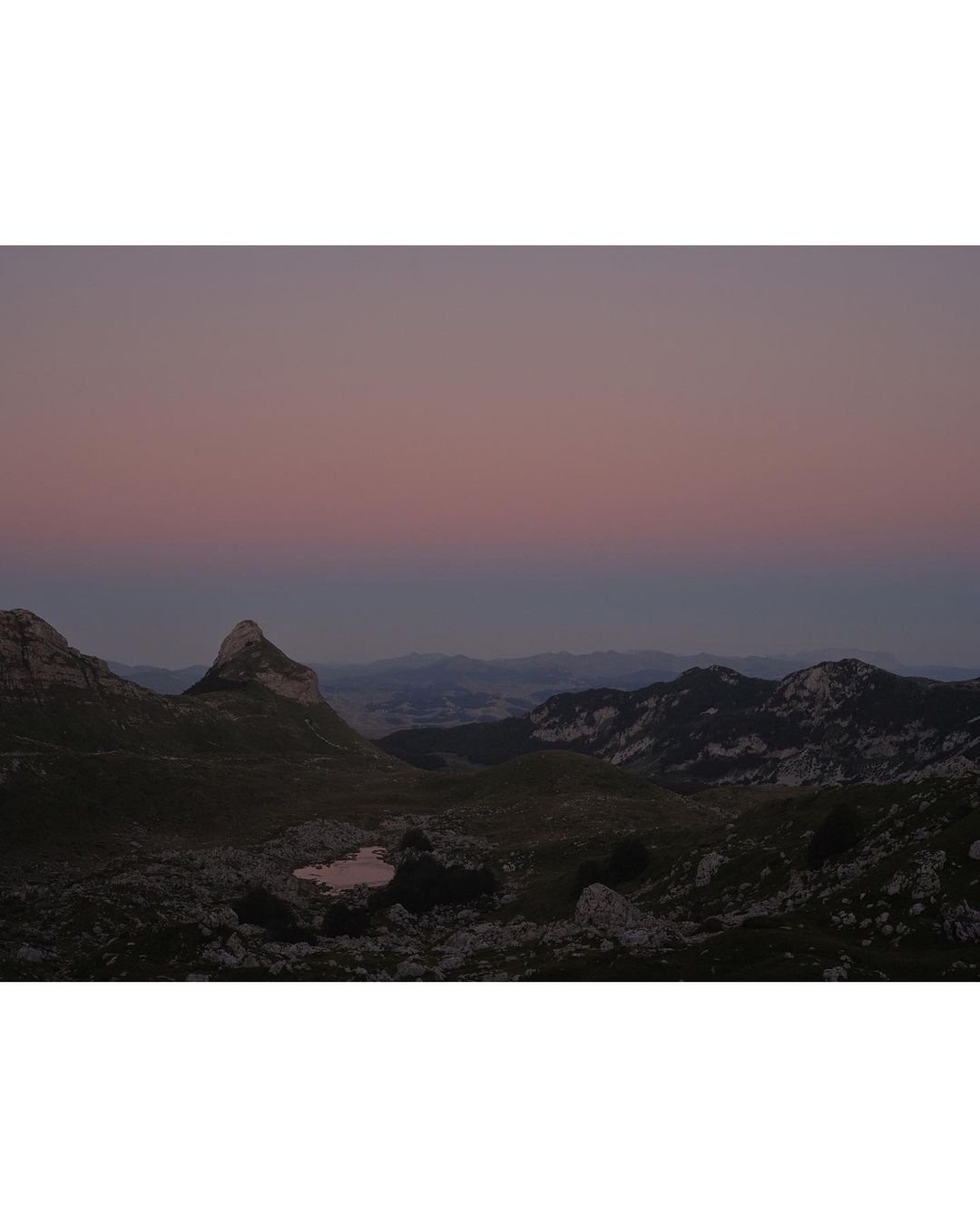 Scenic mountain range landscape view at dusk, featuring rocky terrain, distant peaks, and a soft, gradient sky from blue to pink.