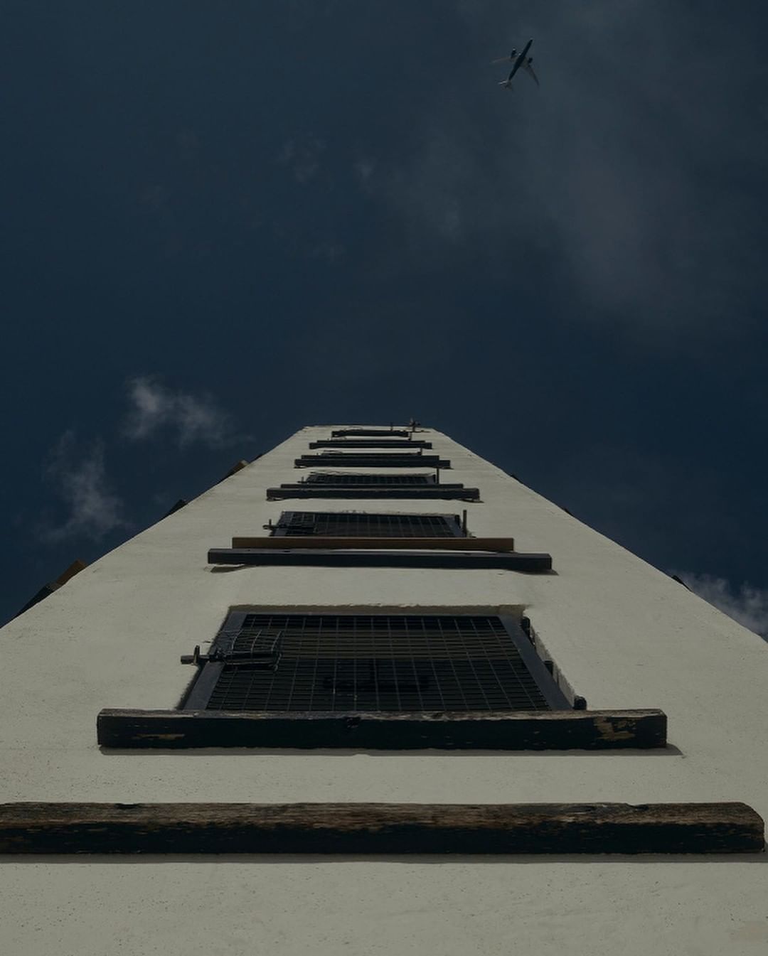 Low-angle view up a white building exterior with multiple windows, leading the eye towards an airplane in the blue sky above.