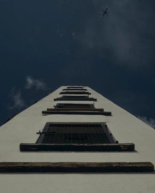 Low-angle view up a white building exterior with multiple windows, leading the eye towards an airplane in the blue sky above.