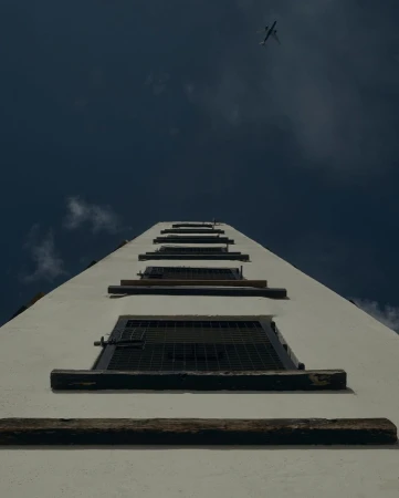 Low-angle view up a white building exterior with multiple windows, leading the eye towards an airplane in the blue sky above.