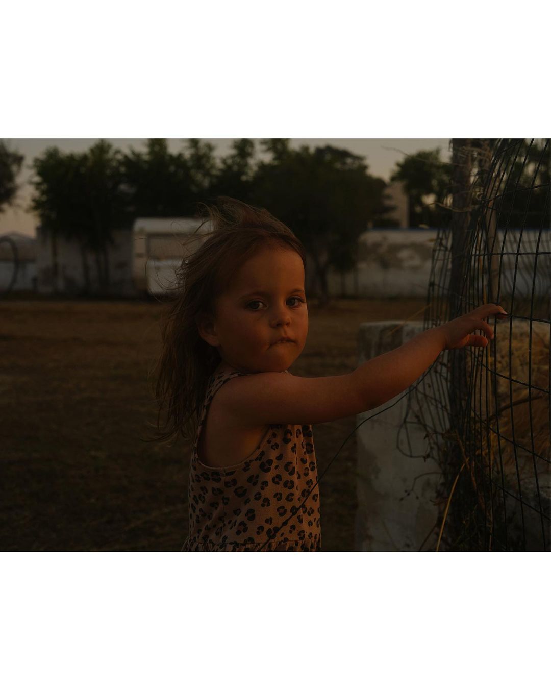 A young child stands near a fence in an outdoor setting, wearing a patterned top with a serious expression, captured in a medium shot.