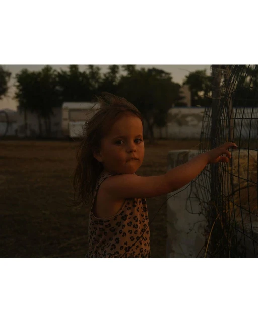 A young child stands near a fence in an outdoor setting, wearing a patterned top with a serious expression, captured in a medium shot.