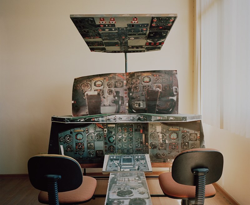 Simulated airplane cockpit set up indoors with chairs in front of the control panel; beige walls and a window with closed blinds.