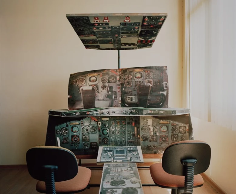 Simulated airplane cockpit set up indoors with chairs in front of the control panel; beige walls and a window with closed blinds.