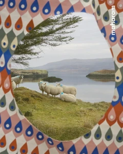 Flock of sheep grazing on a grassy shore by a calm body of water, viewed through a patterned fabric frame