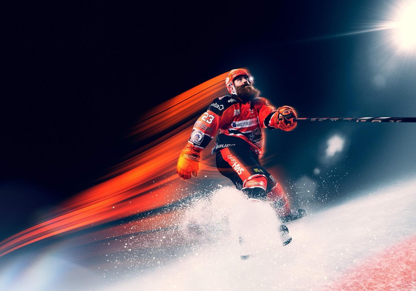 Action shot of a male ice hockey player in red and black uniform with beard gliding on ice, motion blurred in a dynamic sports scene.