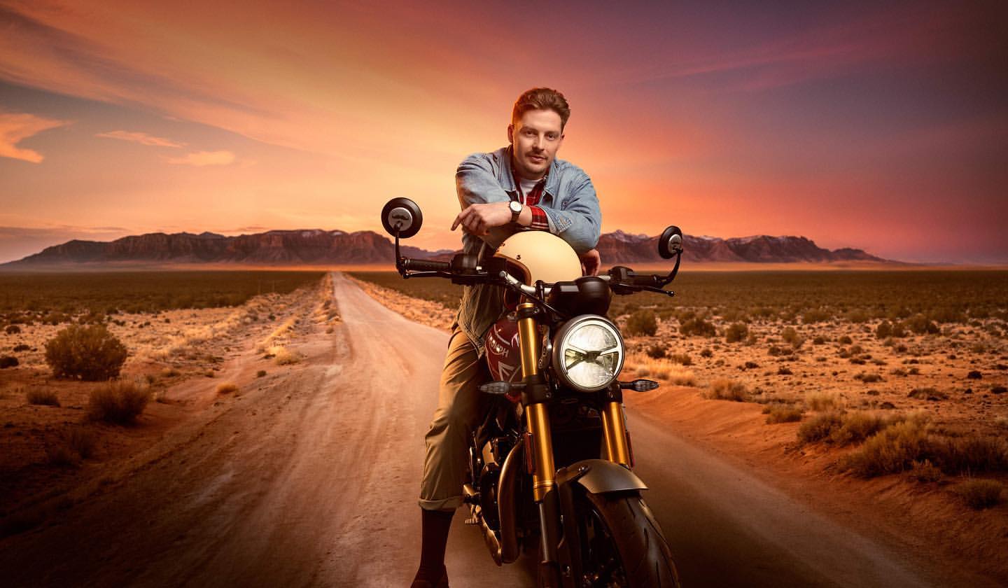 A man sits on a black and gold motorcycle on a desert road at sunset, wearing a denim jacket, with mountains in the background.