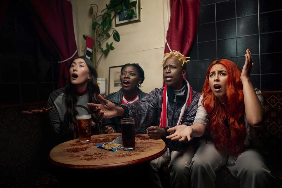 Four diverse adults watch a game in a pub, expressing shock and dismay while seated at a wooden table with drinks and snacks.