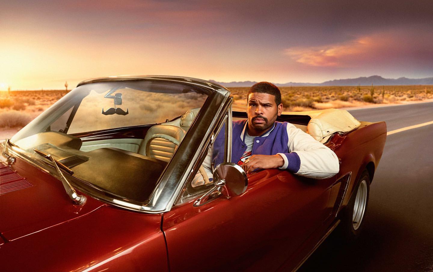 Man in a red convertible car on an open desert road against a sunset sky, wearing a jacket and sporting a mustache air freshener.