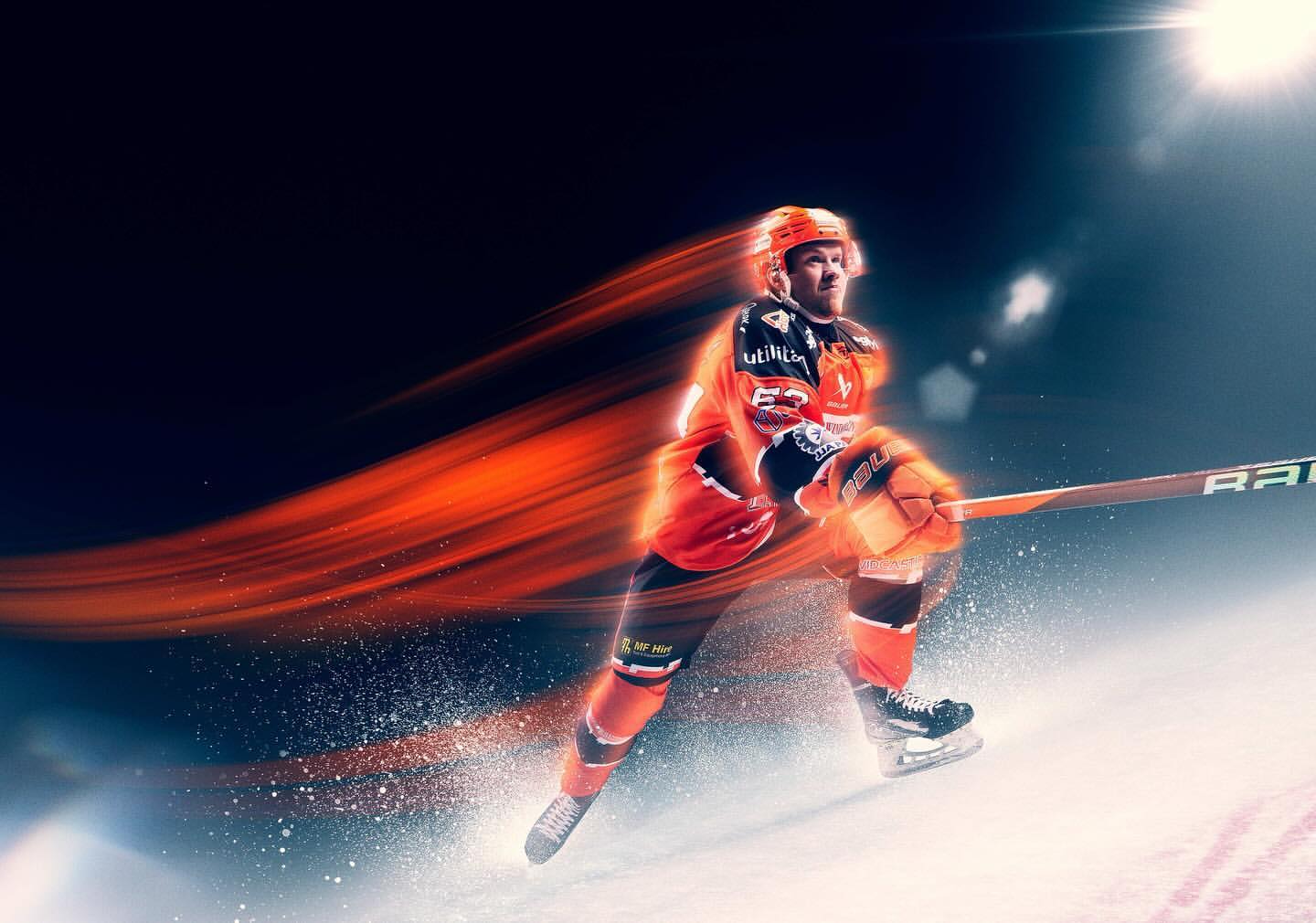 Professional ice hockey player in orange and black uniform skating on the ice rink with motion blur effect and bright spotlight in background.