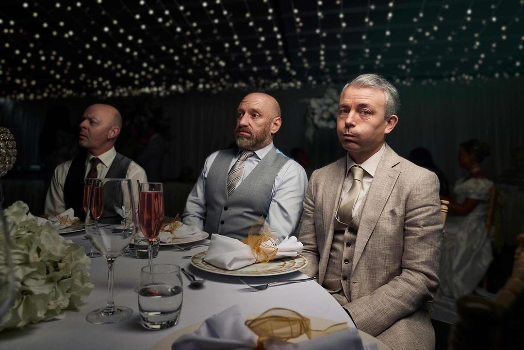 Three adult males seated at a formal table setting in suits at a wedding reception, observed in medium shot, with soft, ambient lighting.