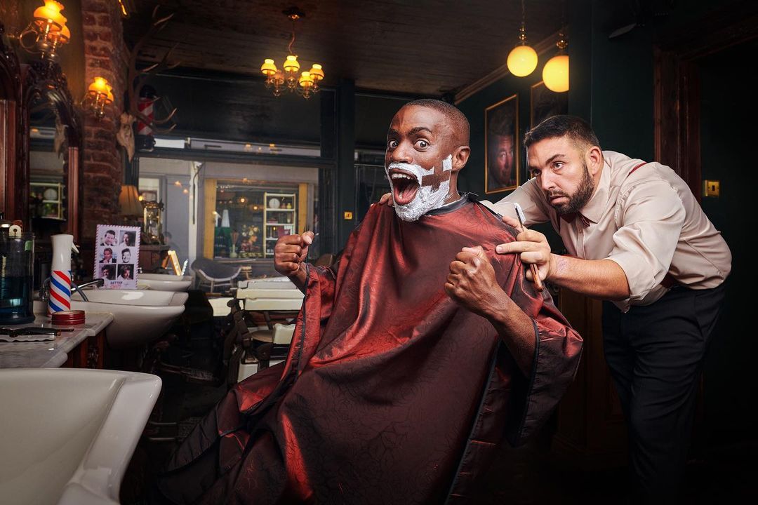 Excited customer with shaving cream on face gets straight razor shave at traditional barber shop with barber; good lighting, classic styling.