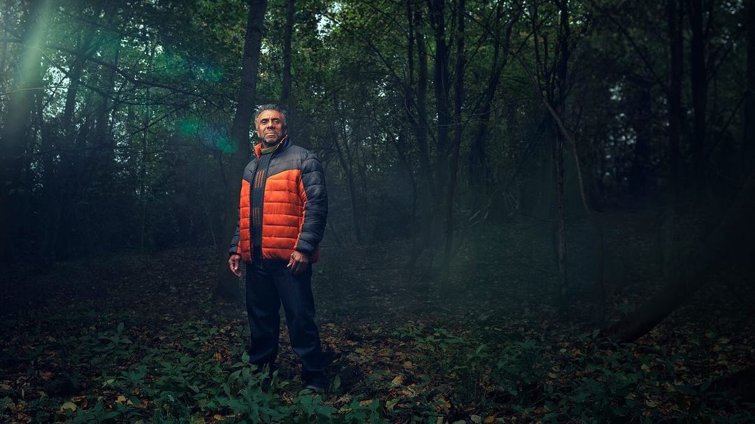 A man stands in a vibrant green forest wearing an orange and grey puffer jacket. The trees are tall, and the light shines through.