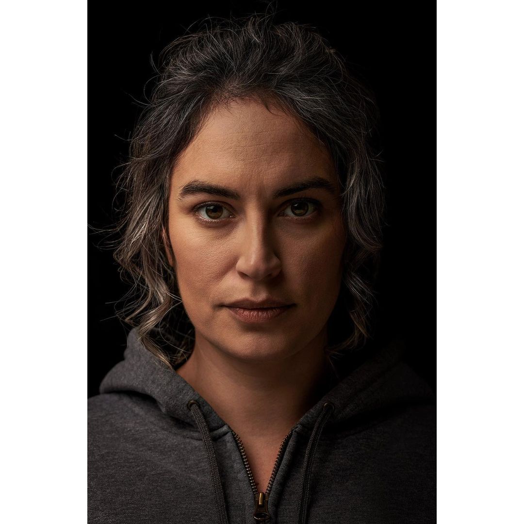 Close-up portrait of an adult woman with graying curly hair in a gray hoodie, set against a dark background with soft, directional lighting.