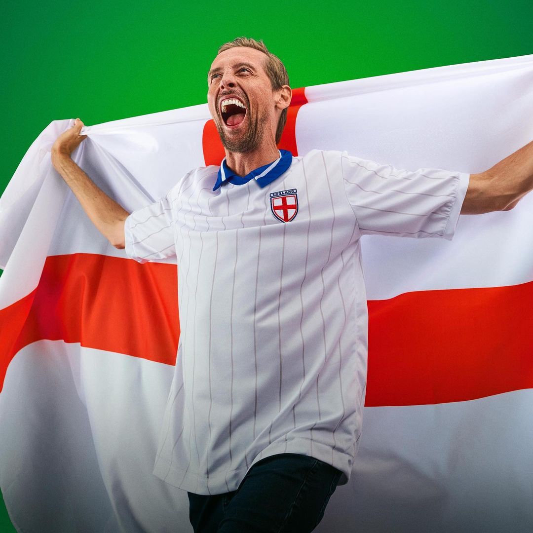 Enthusiastic adult male soccer fan draped in the flag of England, cheering with mouth open wide against a green background.