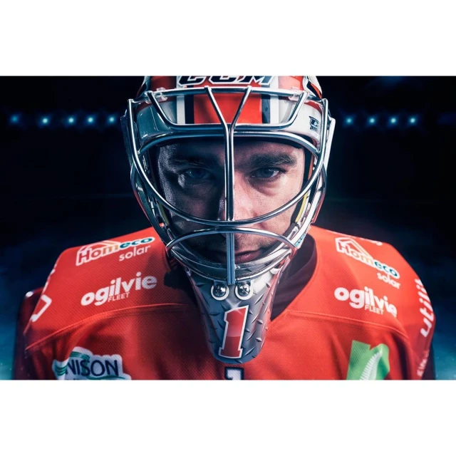 Close-up of an adult male ice hockey goalie wearing a red jersey, mask, and helmet looking directly at the viewer in a dark indoor setting.