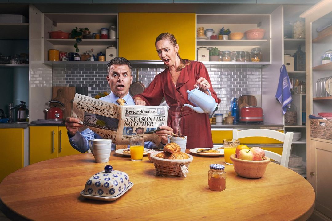 Surprised man reading newspaper as woman pours hot liquid into his mug at breakfast table in a yellow and blue kitchen setting.